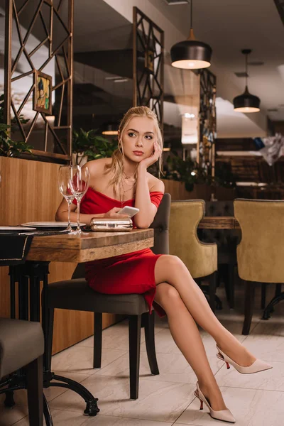 Attractive woman in red dress sitting in restaurant, using smartphone and waiting for man — Stock Photo