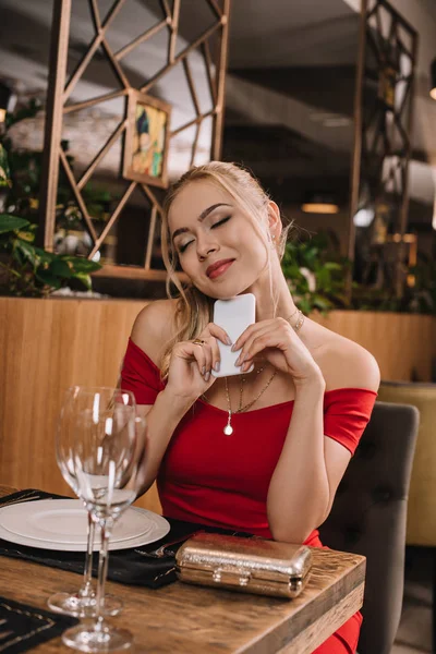 Novia de ensueño en vestido rojo sentado en el restaurante y esperando novio con los ojos cerrados - foto de stock