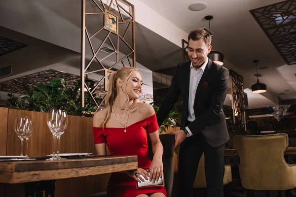 Homem bonito ajudando a sentar mulher atraente em vestido vermelho e segurando cadeira — Fotografia de Stock