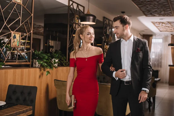 Couple of handsome boyfriend and attractive girlfriend in red dress walking in restaurant and holding hands — Stock Photo