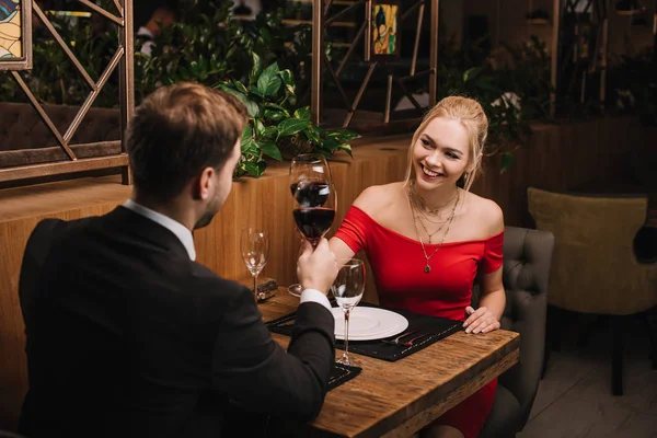 Alegre chica tostando con vino tinto y sonriendo a novio en restaurante - foto de stock