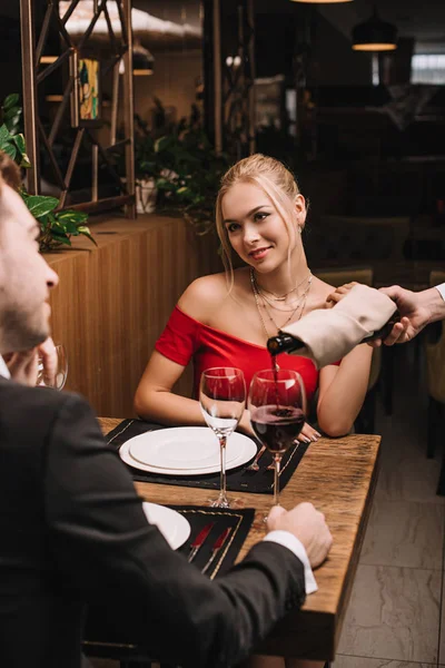 Menina alegre sorrindo para namorado enquanto garçom derramando vinho tinto em vidro no restaurante — Fotografia de Stock