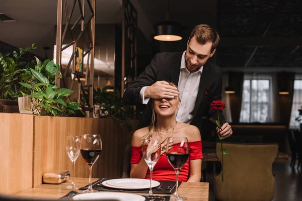 Novio guapo cerrando los ojos de novia atractiva en vestido rojo mientras sostiene rosa - foto de stock