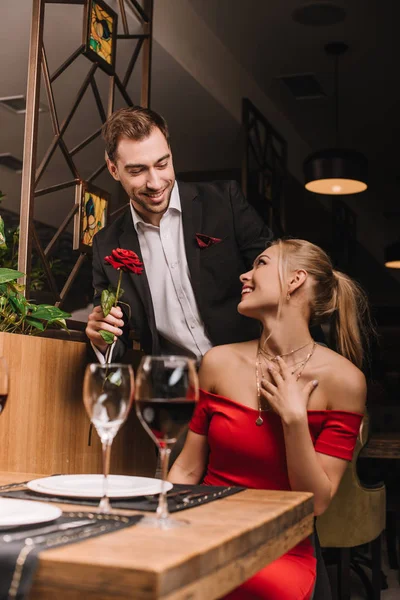 Novio feliz dando rosa roja a atractiva novia en el restaurante - foto de stock