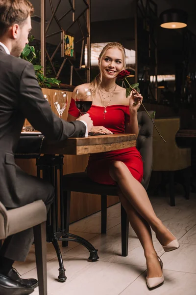 Chica atractiva sentada con rosa roja y mirando novio en el restaurante - foto de stock