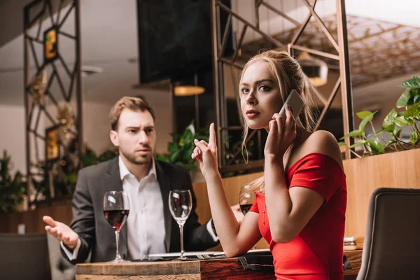 Hombre insatisfecho mirando a su novia hablando en el teléfono inteligente durante la cena - foto de stock
