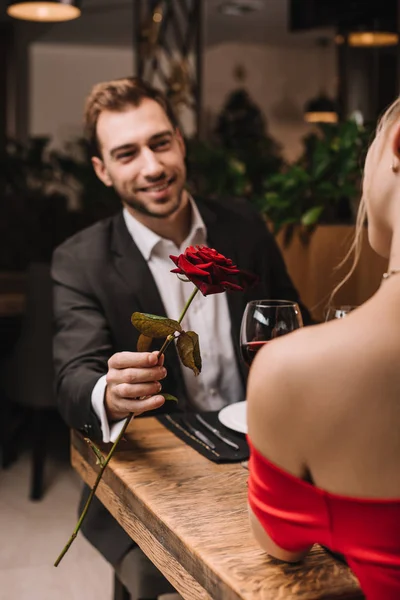 Selective focus of red rose in hand of happy man looking at girlfriend in restaurant — Stock Photo