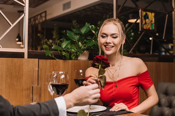 Messa a fuoco selettiva di fidanzata allegra guardando rosa rossa in mano del fidanzato mentre si siede nel ristorante — Foto stock