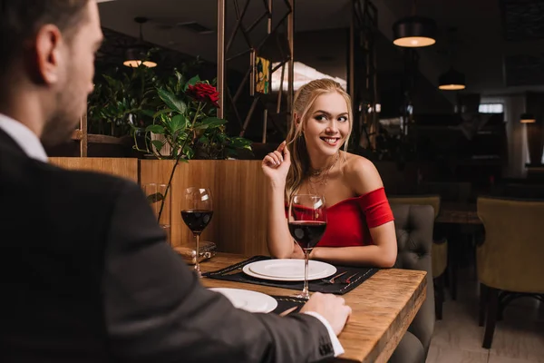 Atractiva mujer coqueteando con su novio mientras está sentado en vestido rojo en el restaurante - foto de stock