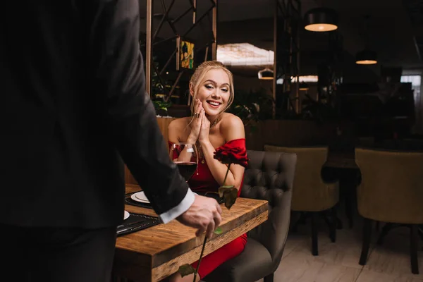 Chica feliz mirando rosa roja en la mano del hombre mientras está sentado en el restaurante - foto de stock