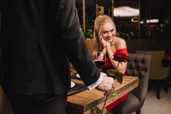 Selective focus of happy woman looking at red rose in hand of man and smiling while sitting in restaurant — Stock Photo