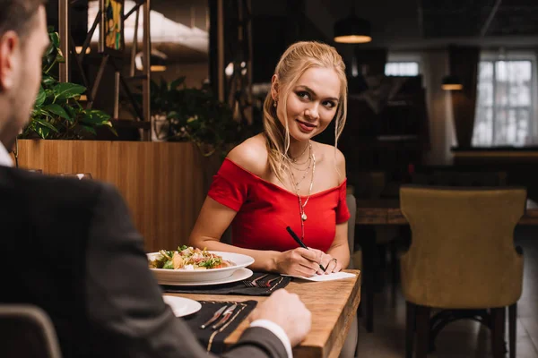 Attraktive blonde Frau schreibt auf Papier, während sie ihren Freund im Restaurant ansieht — Stockfoto