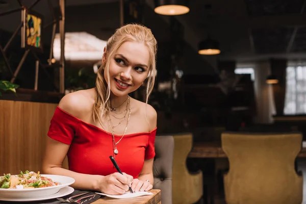 Fröhliche blonde Frau, die auf Papier schreibt und im Restaurant lächelt — Stockfoto