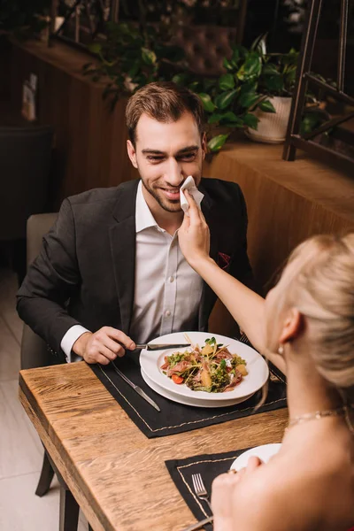 Fidanzata asciugare la bocca del fidanzato allegro con tovagliolo nel ristorante — Foto stock