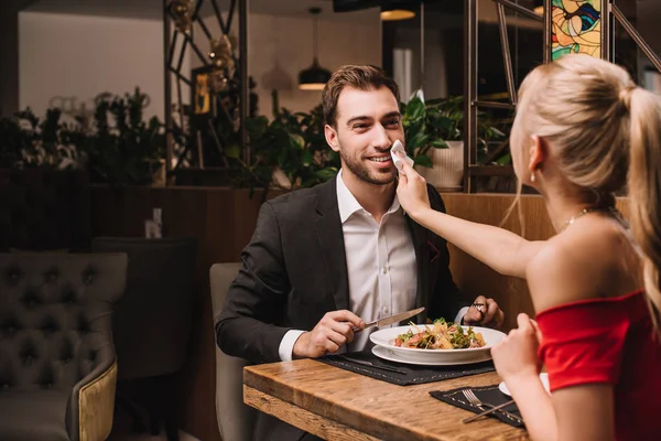 Mujer limpiando la boca de feliz novio con servilleta en el restaurante - foto de stock