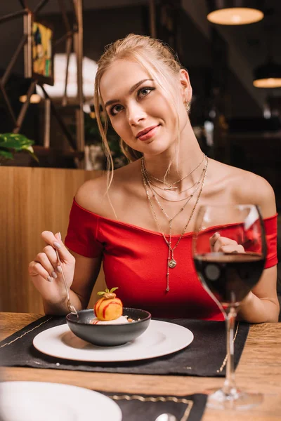 Dreamy girl sitting in red dress and holding spoon near sweet dessert in restaurant — Stock Photo