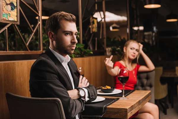 Selective focus of offended man sitting with crossed arms while girlfriend showing middle finger during dinner — Stock Photo