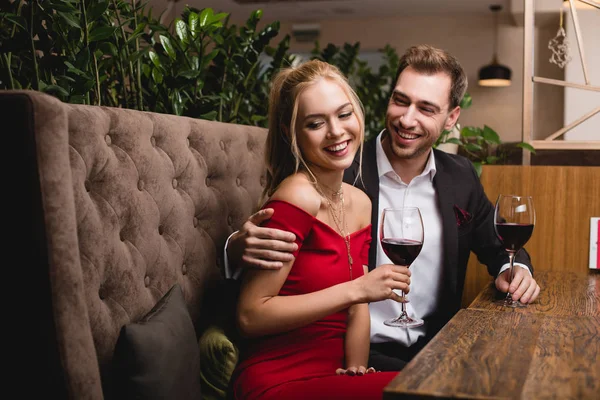 Menina feliz rindo perto namorado enquanto segurando vidro com vinho tinto no restaurante — Fotografia de Stock