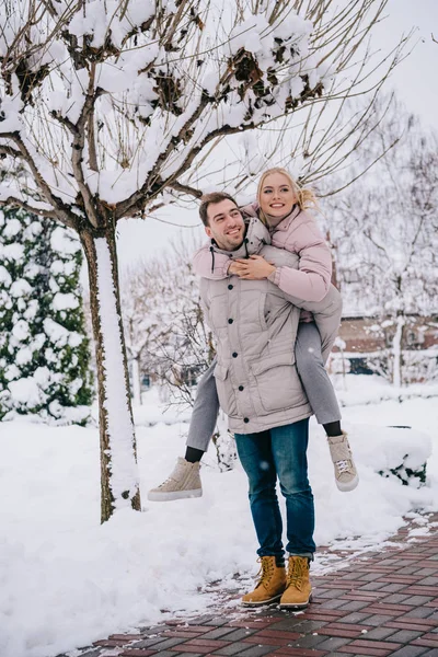 Homem alegre piggybacking sorrindo namorada na rua nevada — Fotografia de Stock