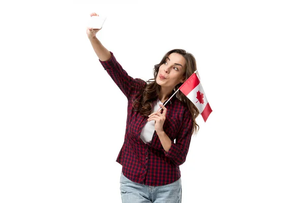 Женщина с канадским флагом, высовывая язык и делая селфи на смартфоне, изолированном на белом — стоковое фото