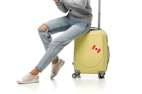 Vista recortada de la mujer sentada en la maleta con pegatina de bandera canadiense aislado en blanco, concepto de viaje - foto de stock