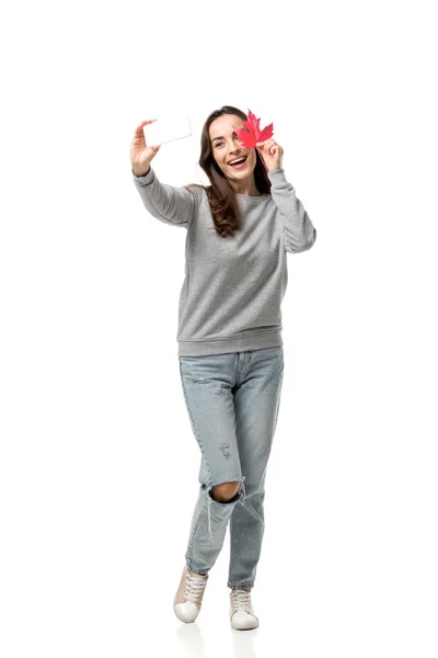 Happy woman in casual clothes holding maple leaf and taking selfie on smartphone isolated on white — Stock Photo