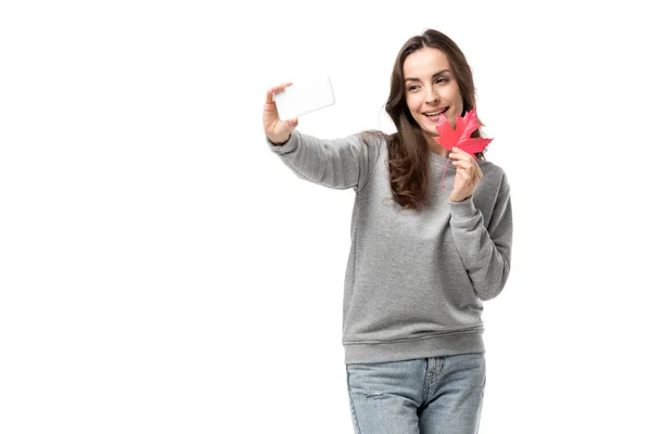 Mujer sonriente en ropa casual sosteniendo hoja de arce y tomando selfie en el teléfono inteligente aislado en blanco - foto de stock