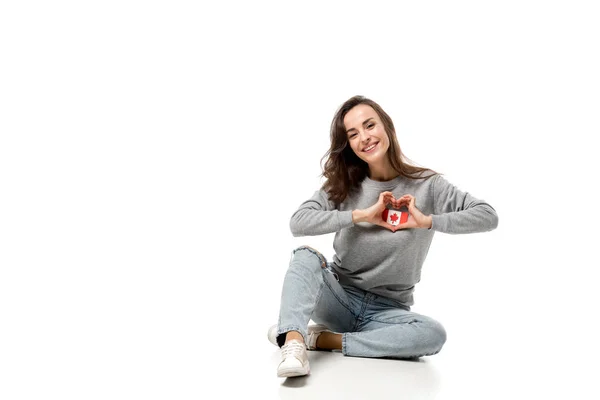 Woman sitting and showing heart symbol with hands over canadian flag badge isolated on white — Stock Photo