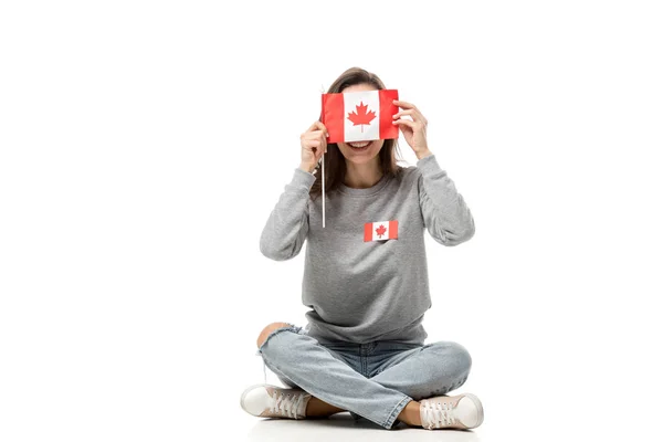 Femme avec insigne de feuille d'érable assise et tenant le drapeau canadien devant le visage isolé sur blanc — Photo de stock