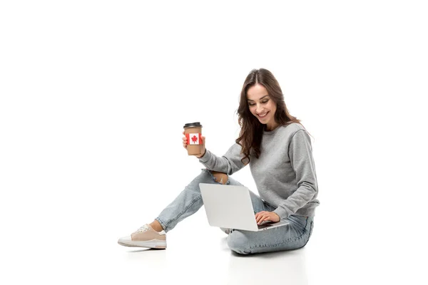 Mujer sonriente usando el ordenador portátil y sosteniendo la taza de café con etiqueta engomada bandera canadiense aislado en blanco - foto de stock