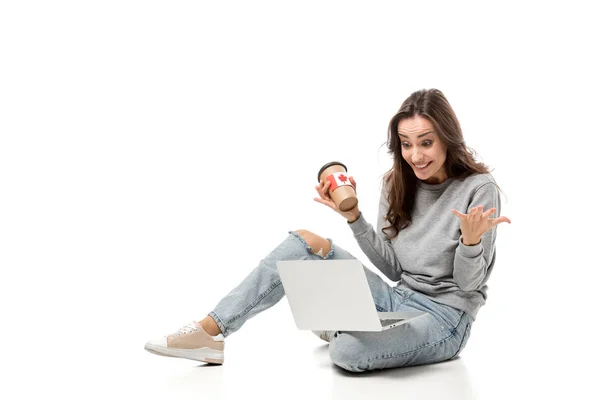 Mujer sorprendida usando el ordenador portátil y sosteniendo la taza de café con etiqueta engomada bandera canadiense aislado en blanco - foto de stock