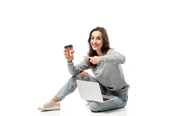 Mujer feliz usando el ordenador portátil y apuntando con el dedo a la taza de café con pegatina de bandera canadiense aislado en blanco - foto de stock