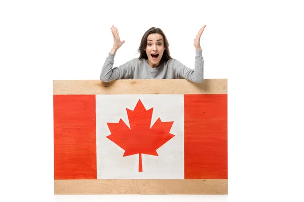 Donna sorpresa gesticolando con le mani dietro tavola di legno con bandiera canadese isolata su bianco — Foto stock