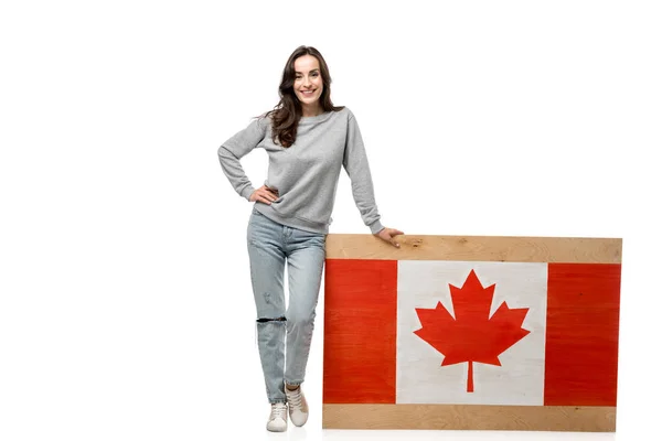 Smiling woman with hand on hip standing near wooden board with canadian flag isolated on white — Stock Photo