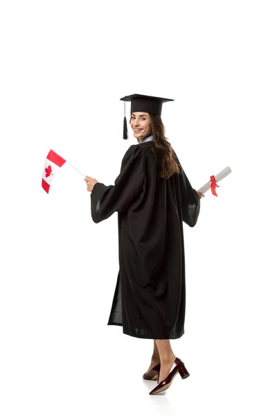 Sonriente estudiante con bata académica sosteniendo bandera canadiense y diploma aislado en blanco - foto de stock