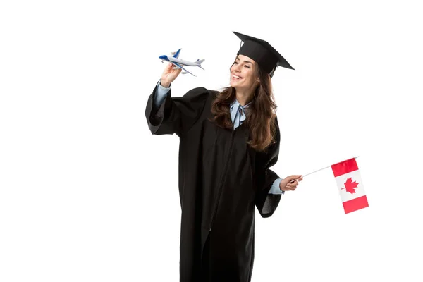Щаслива студентка в академічній сукні тримає канадський прапор і модель літака ізольована на білому, навчається за кордоном концепція — стокове фото