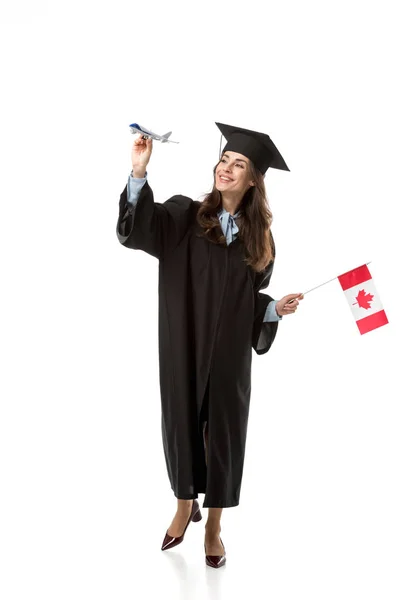 Smiling female student in academic gown holding canadian flag and plane model isolated on white, studying abroad concept — Stock Photo