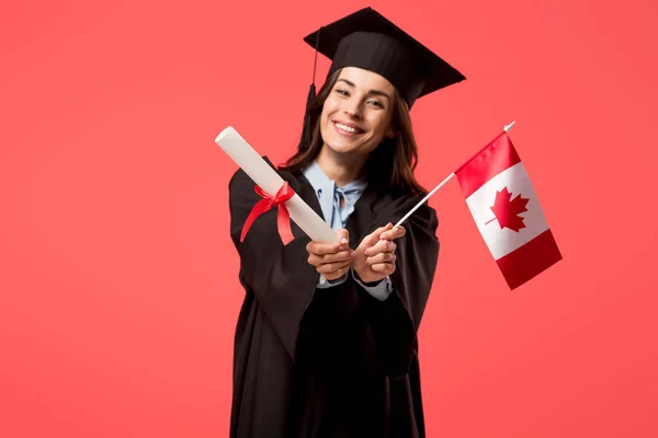 Sonriente estudiante en vestido académico sosteniendo bandera canadiense aislado en coral vivo - foto de stock