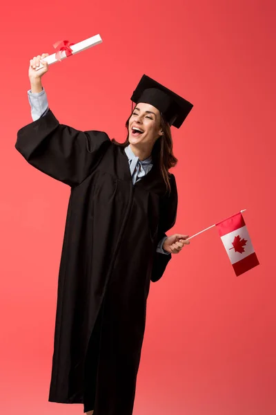 Feliz estudiante en vestido académico sosteniendo bandera canadiense y diploma aislado en coral vivo - foto de stock