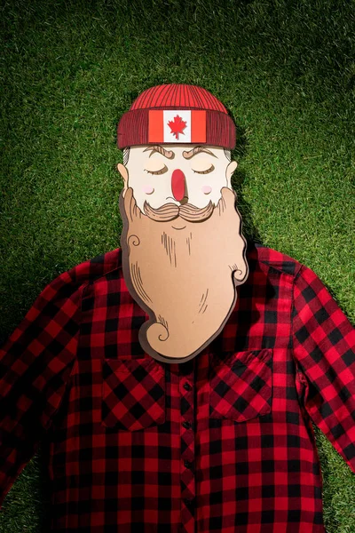 Cardboard man in plaid shirt and hat with maple leaf on green grass background — Stock Photo
