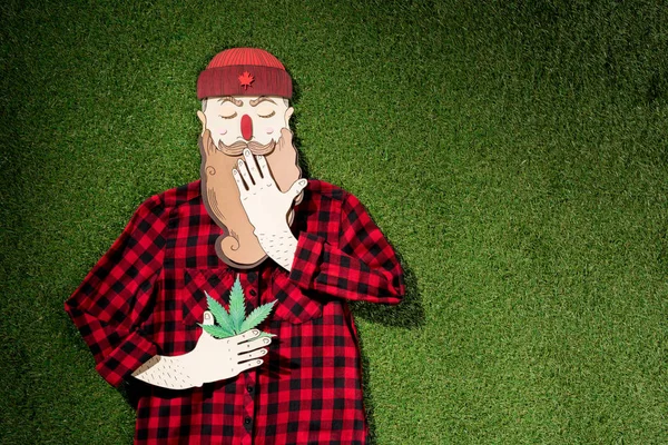 Homme en carton en chemise à carreaux tenant du cannabis et couvrant la bouche avec la main sur fond d'herbe verte, concept de légalisation de la marijuana — Photo de stock