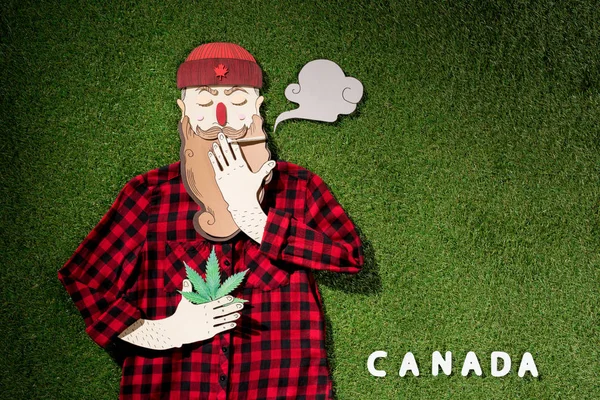 Vista superior del hombre de cartón con camisa a cuadros sosteniendo cannabis y fumando sobre hierba verde de fondo, concepto de legalización de la marihuana - foto de stock