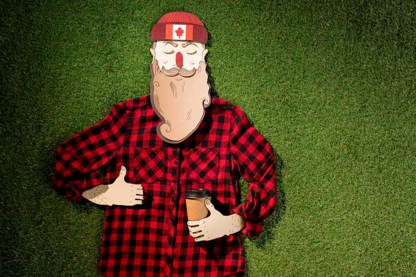 Cardboard man in plaid shirt holding coffee and showing thumb up sign on green grass background — Stock Photo