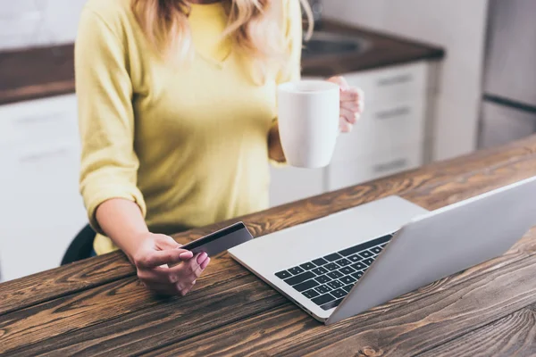 Vista recortada de la mujer sosteniendo la taza con la bebida y la tarjeta de crédito cerca del ordenador portátil - foto de stock