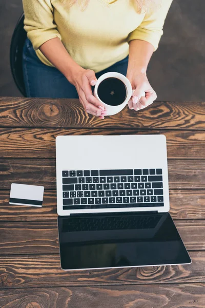 Обрезанный вид женщины, держащей чашку кофе возле ноутбука и кредитной карты — стоковое фото
