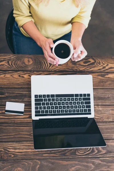 Cropped view of woman holding cup of coffee near laptop and credit card — Stock Photo