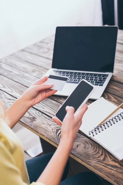 Cropped view of woman holding smartphone and credit card near laptop with blank screen — Stock Photo