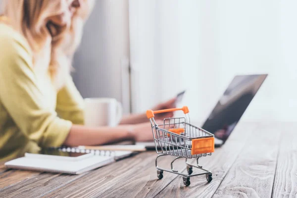 Selective focus of small shopping trolley with woman using laptop on background — Stock Photo