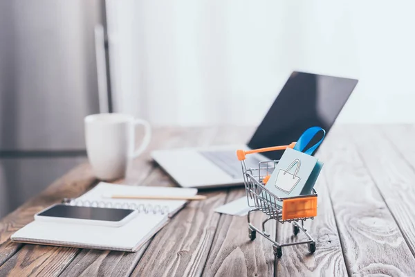 Foco seletivo do saco de compras de brinquedos no carrinho de compras pequeno com laptop em segundo plano — Fotografia de Stock