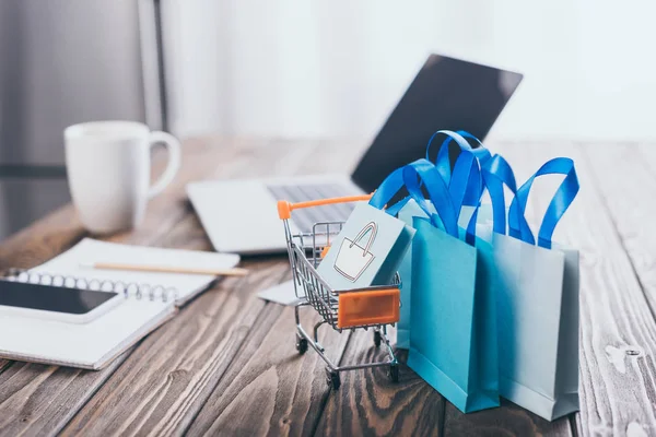 Selective focus of small shopping bags near toy shopping trolley with laptop on background — Stock Photo